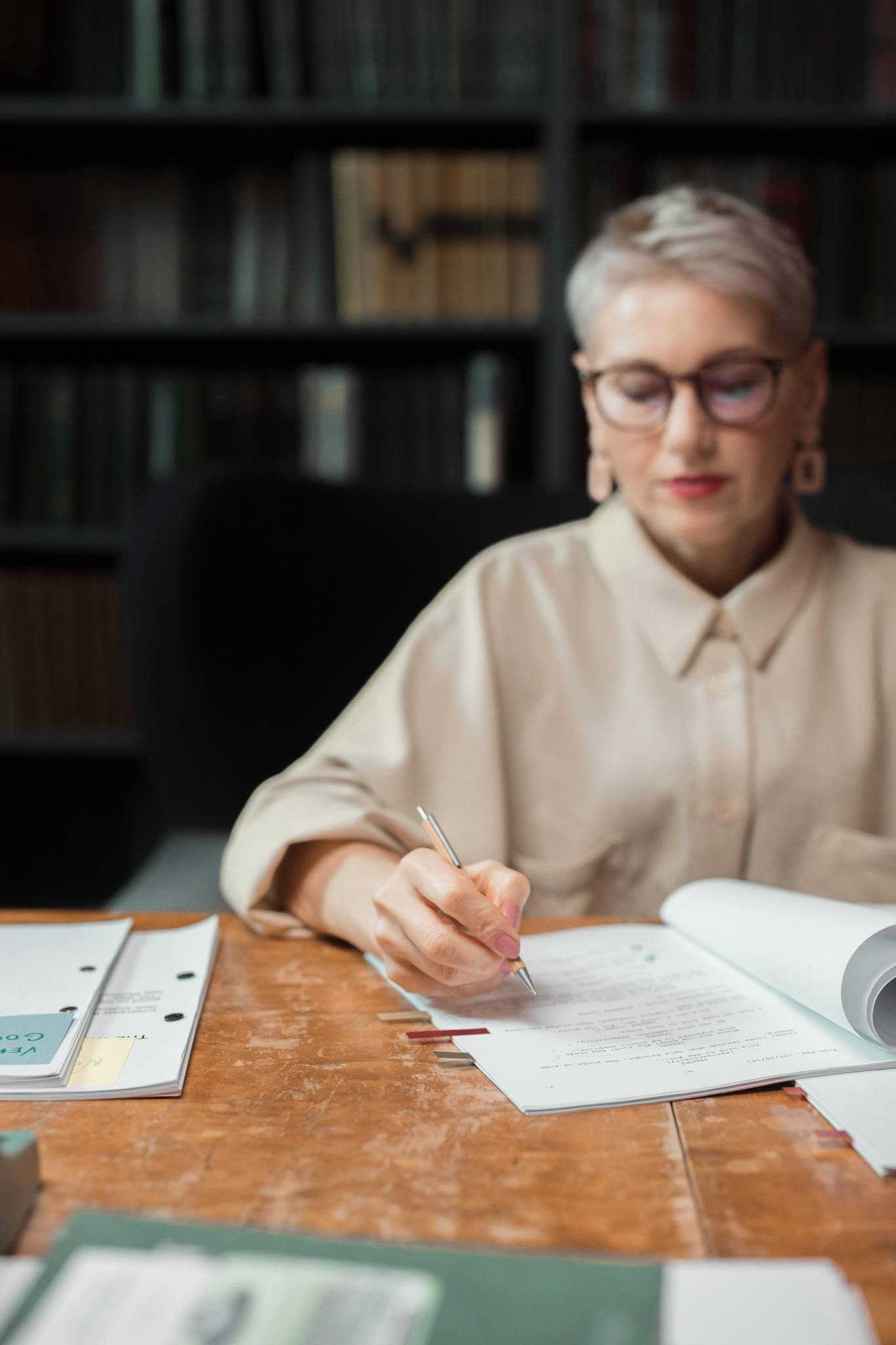 Elderly woman in glasses writing a manuscript at a desk in a library setting, focused on her work.